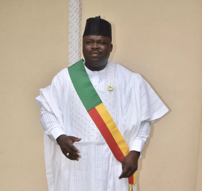 Homme en tenue traditionnelle blanche avec écharpe tricolore vert, jaune et rouge et chapeau noir sur fond beige