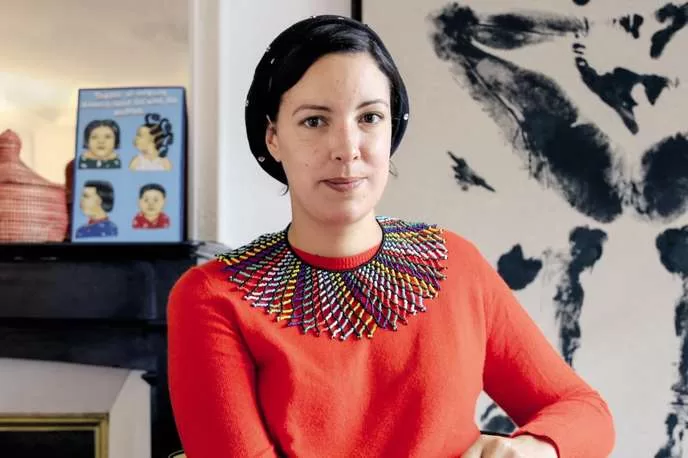 Femme portant un pull rouge et un collier coloré assise devant un mur décoré de peintures et d'objets d'art