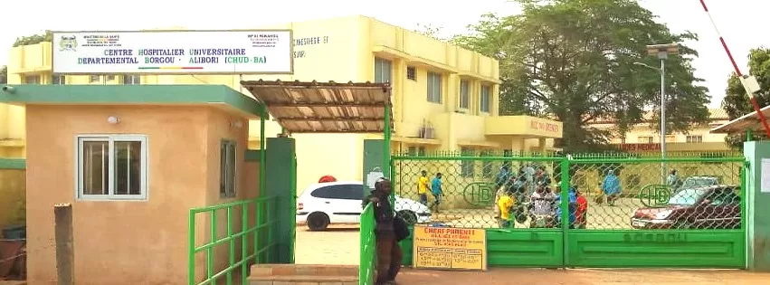 Entrée d un centre hospitalier avec portail vert et plusieurs personnes à l intérieur et à l’extérieur du site en journée