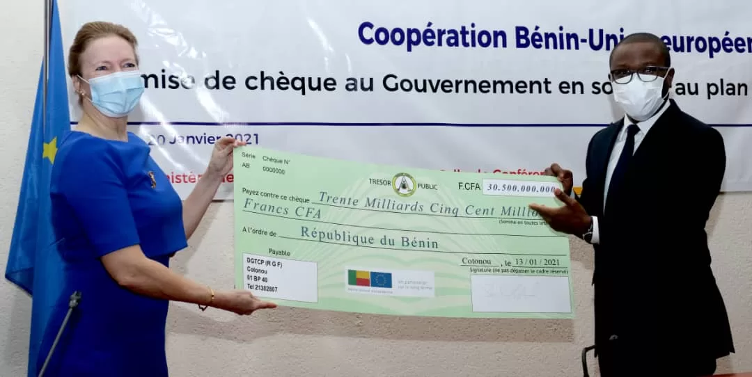 Deux personnes masquées tiennent un grand chèque représentant un don financier officiel devant une bannière de coopération internationale