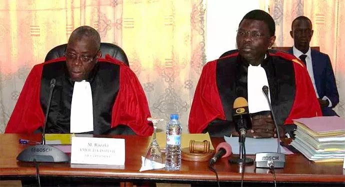 Deux hommes en toges rouges et blanches assis à une table avec des microphones et des dossiers devant eux