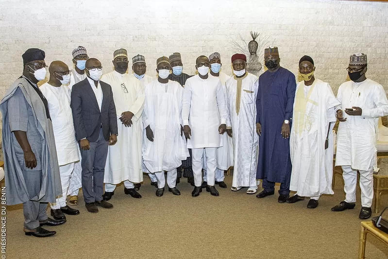 Groupe d’hommes en tenue traditionnelle africaine et costume occidental posant ensemble dans une pièce avec un mur en pierre claire