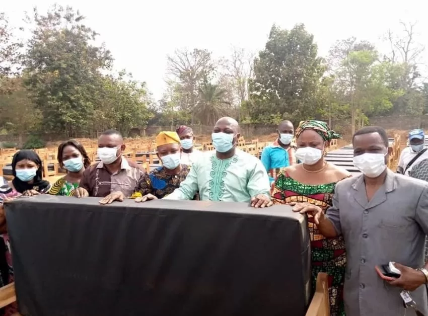 Un groupe de personnes masquées tenant un lit avec matelas devant de nombreux autres lits alignés en extérieur avec des arbres en arrière-plan