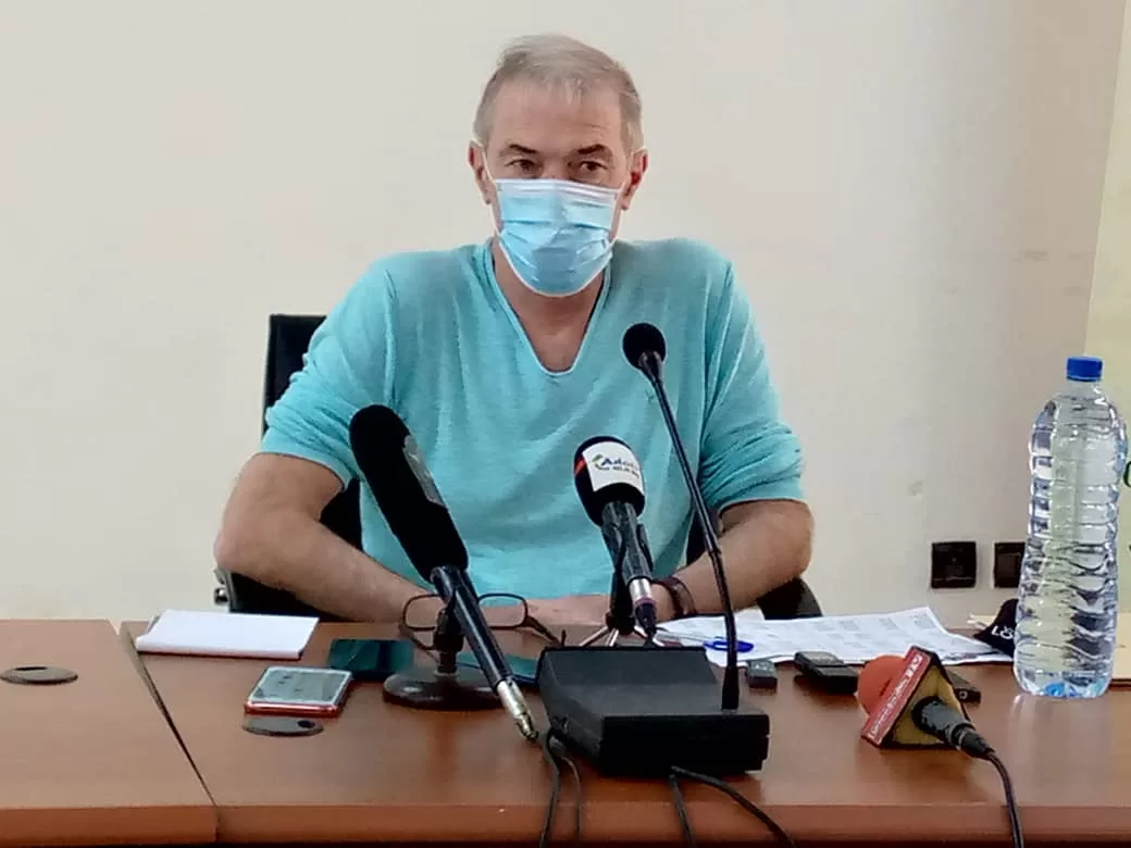 Homme masqué en chemise bleue assis derrière une table avec plusieurs micros et une grande bouteille d'eau