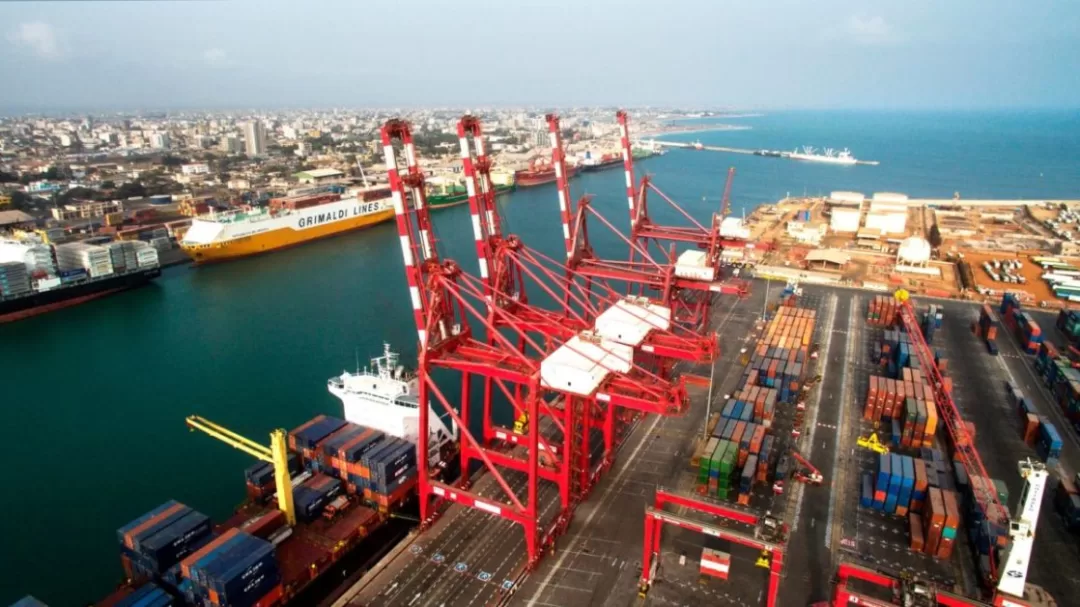 Grand port maritime avec plusieurs grues rouges et conteneurs empilés le long du quai face à la mer et la ville en arrière-plan