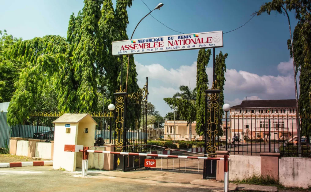 Entrée principale d un bâtiment officiel entouré de plantations avec un portail métallique fermé et une barrière rouge et blanche