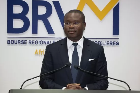 Homme en costume sombre et cravate debout derrière un pupitre avec microphones devant un logo BRVM sur un mur blanc