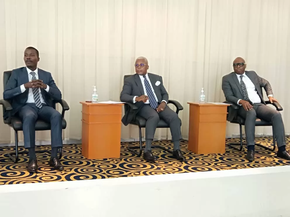Trois hommes en costume assis sur des fauteuils dans une salle avec tapis à motifs et murs clairs