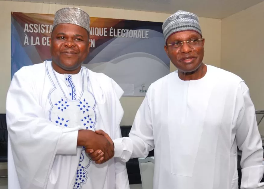 Deux hommes en tenue traditionnelle blanche se serrent la main devant une bannière partiellement visible sur fond bleu