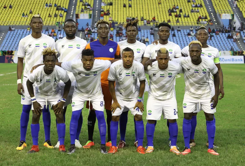Onze joueurs de football en tenue blanche posent sur un terrain avant un match dans un stade avec des gradins jaunes et bleus