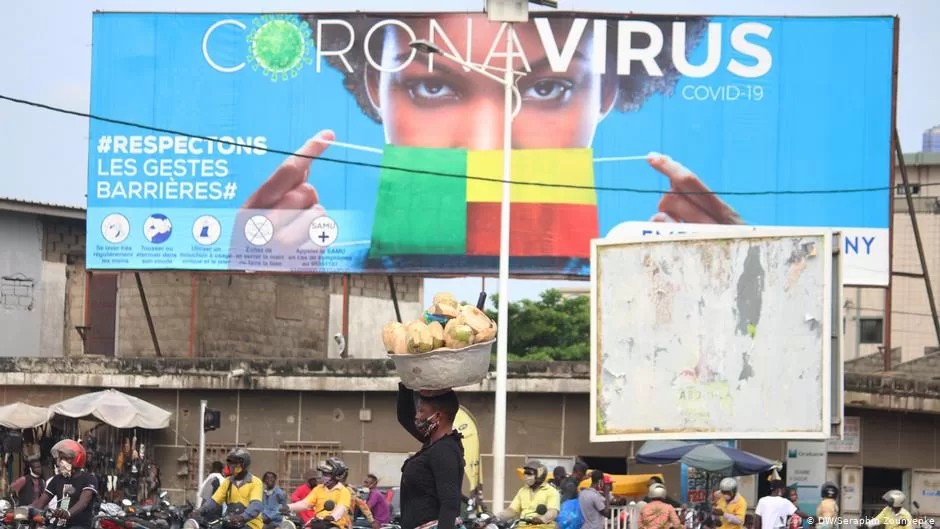 Foule dense devant un grand panneau sur le coronavirus avec un visage masqué aux couleurs d’un drapeau et une femme portant un panier sur la tête