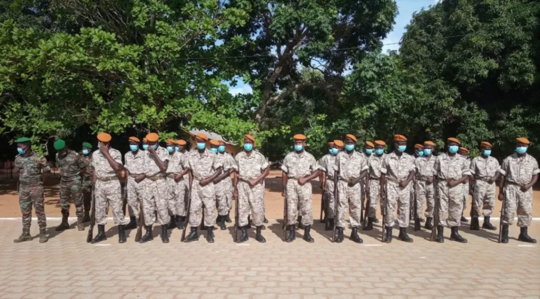 Un groupe de militaires en uniforme clair et portant des masques, alignés en rangs dans un espace extérieur arboré