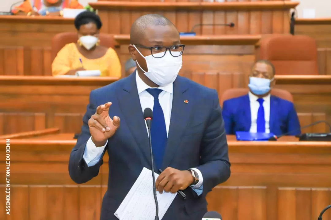 Homme en costume cravate et masque parlant debout dans une salle d’assemblée avec des personnes assises en arrière-plan