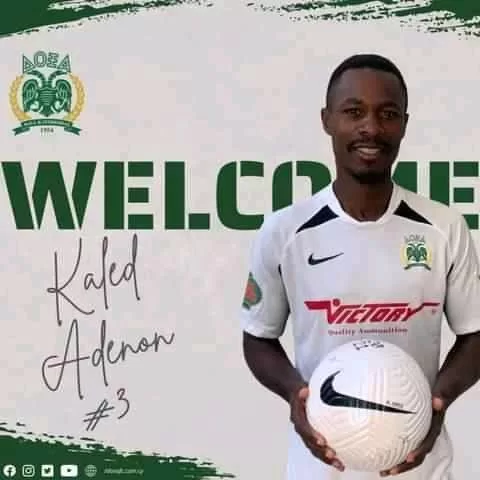 Un homme souriant en maillot de football blanc tient un ballon devant un mur blanc affichant un message de bienvenue en vert