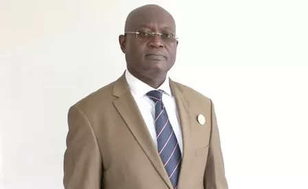 Homme en costume marron clair, cravate rayée, portant des lunettes, posé devant un mur blanc