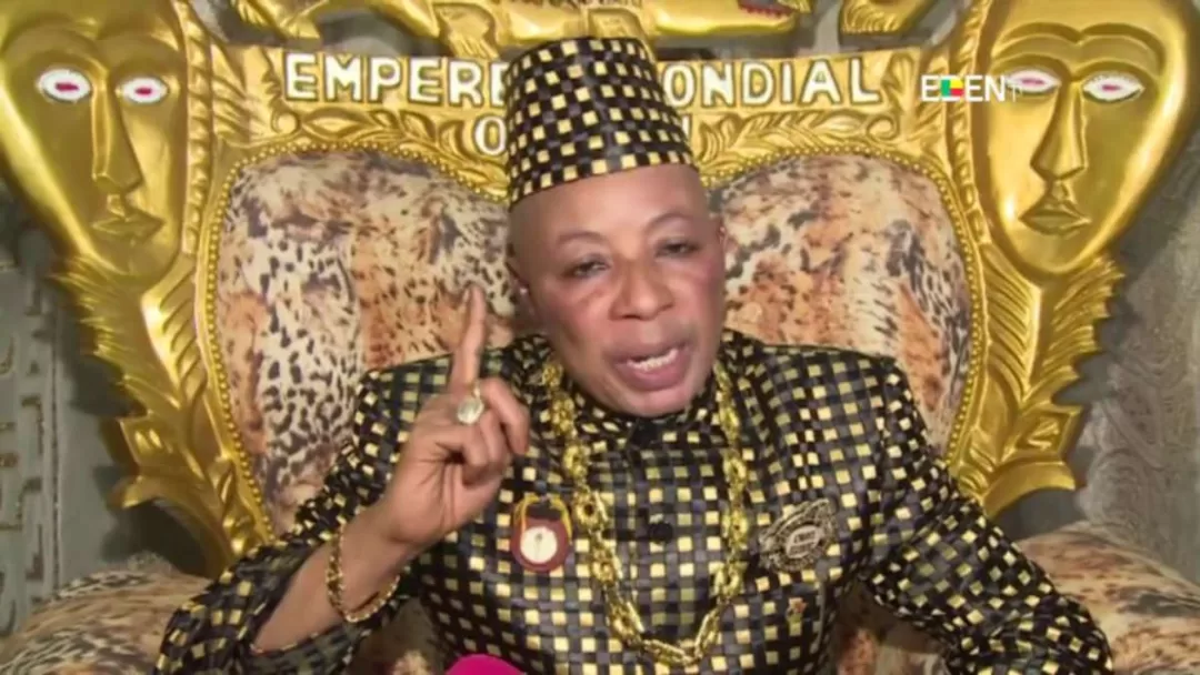 Un homme en tenue traditionnelle à motifs noirs et jaunes, assis sur un fauteuil doré orné de visages, parlant et levant un doigt