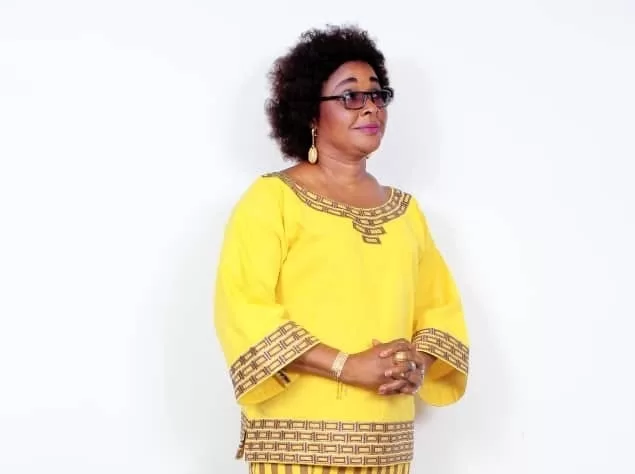 Femme debout, vêtue d’une robe jaune ornée de motifs marron, les mains jointes et regard tourné vers la droite