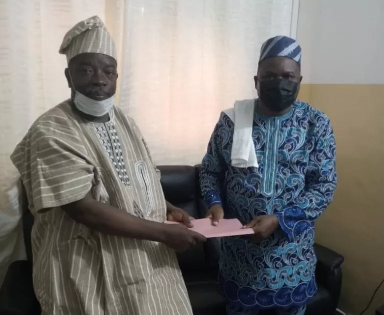 Deux hommes en tenue traditionnelle et masque tenant un document rose dans une pièce avec un canapé noir et un rideau beige