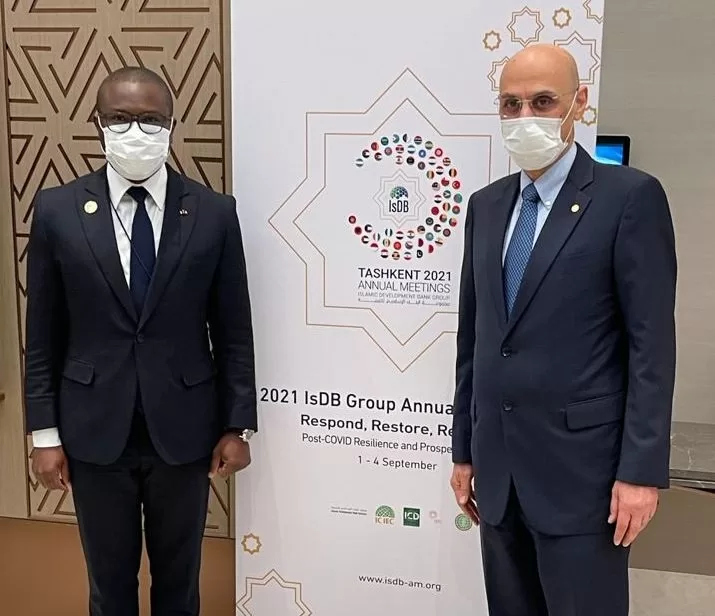 Deux hommes en costume sombre et masque chirurgical posent devant une bannière d'une conférence internationale