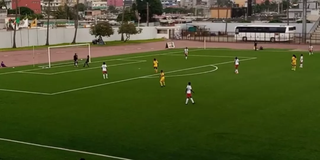 Dix joueuses en action sur un terrain de football vert entouré de bâtiments urbains et d’un bus blanc stationné à l’arrière-plan