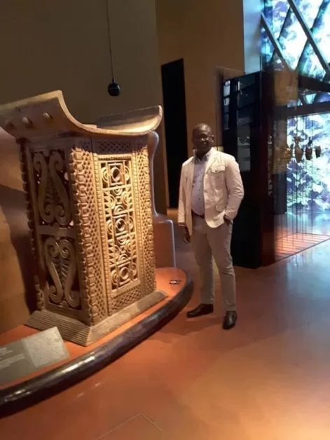Un homme en costume clair pose près d une grande sculpture ou objet décoratif en bois sculpté dans une salle d exposition lumineuse