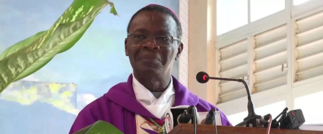 Un homme en tenue religieuse violette parle derrière un pupitre avec micro dans une pièce éclairée par une fenêtre blanche