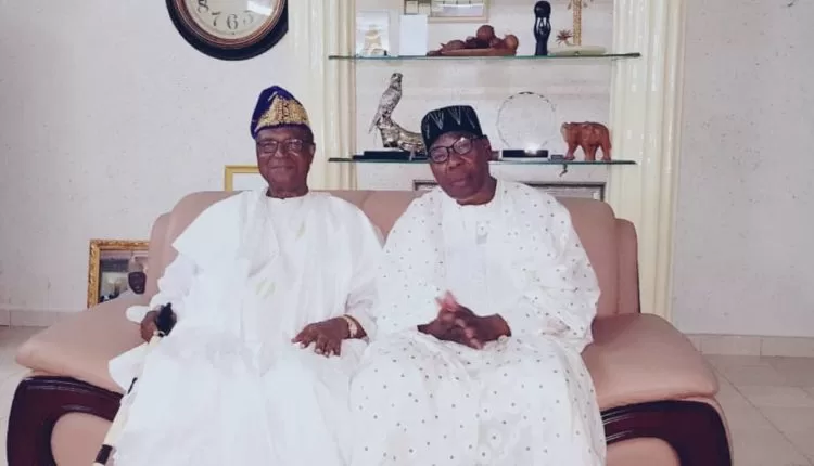 Deux hommes âgés en tenue traditionnelle africaine blanche assis sur un canapé devant une vitrine avec objets décoratifs