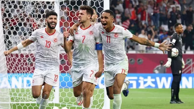 Trois joueurs en maillots blancs célèbrent un but devant le but avec le gardien adverse en arrière-plan.