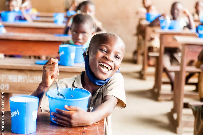 Un enfant souriant mange dans une cantine scolaire avec d’autres enfants assis à des pupitres en arrière-plan