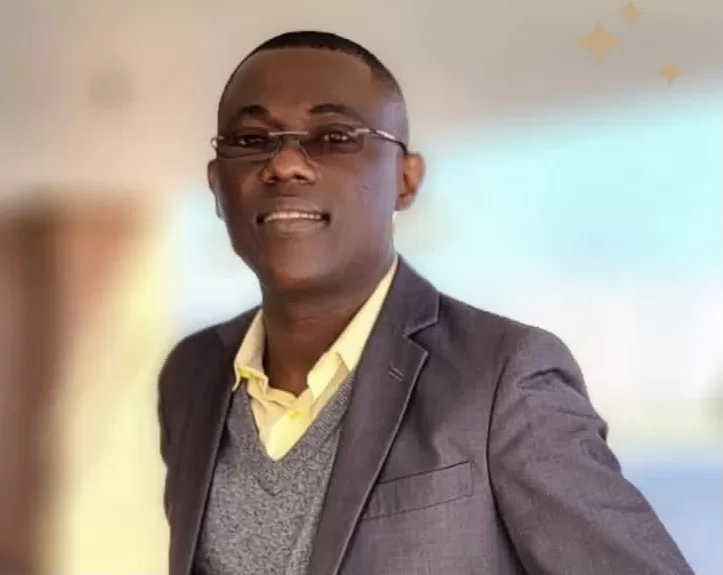 Homme portant des lunettes, un costume sombre avec un gilet gris et une chemise jaune souriant devant un fond flou