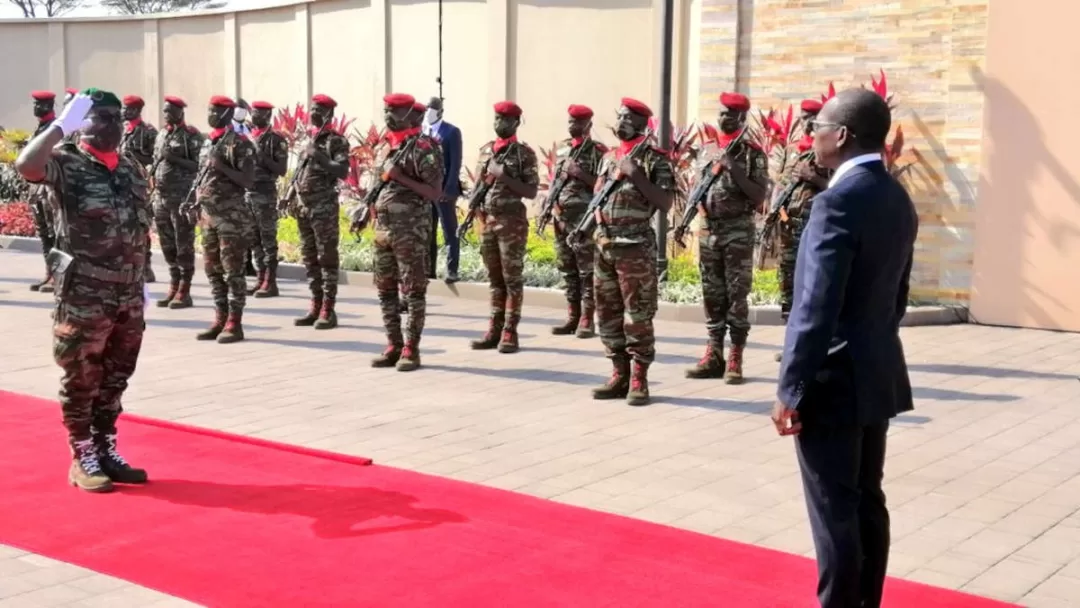 Un groupe de soldats en tenue camouflée et bérets rouges en formation face à un homme en costume sur un tapis rouge devant un mur beige