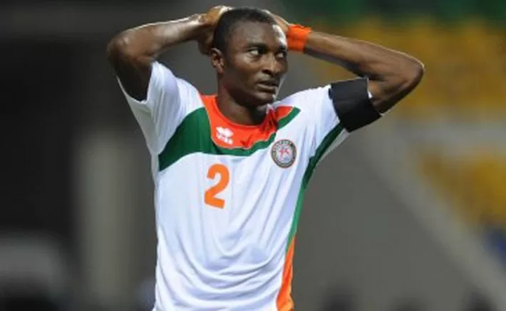 Joueur de football en maillot blanc aux bandes vertes et oranges les mains sur la tête expression contrariée sur le terrain