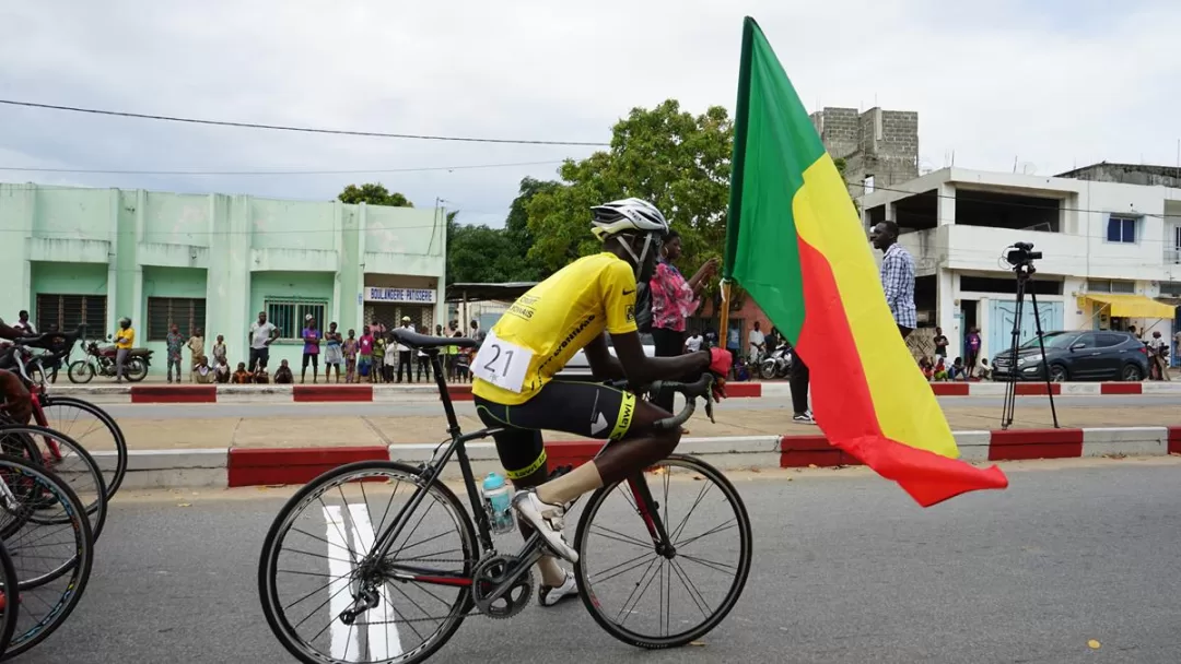 Un cycliste en tenue jaune et casque porte un drapeau tricolore rouge, jaune et vert lors d'une course en ville