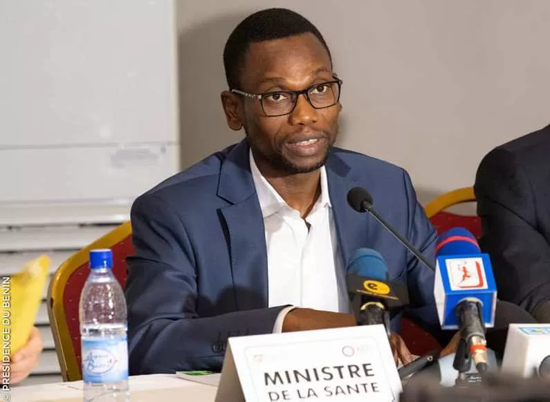 Un homme en costume bleu parle au micro lors d'une conférence avec une pancarte "MINISTRE DE LA SANTE" devant lui