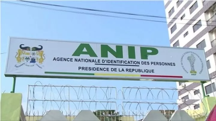 Panneau avec le sigle ANIP Agence Nationale d’Identification des Personnes au-dessus d’une clôture et de barbelés devant un immeuble.