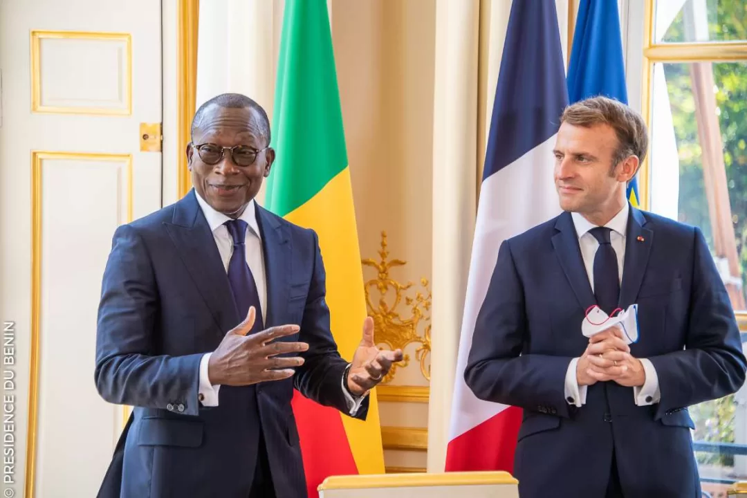 Deux hommes en costume discutent devant deux drapeaux dont un tricolore français dans une salle élégante avec une grande fenêtre