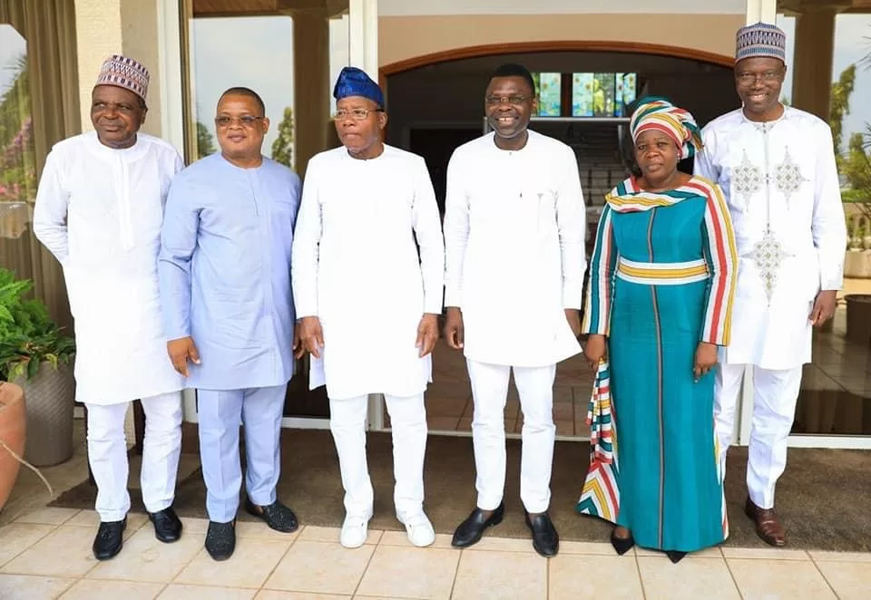 Six personnes debout côte à côte devant une porte vitrée, portant des tenues traditionnelles et formelles colorées variées