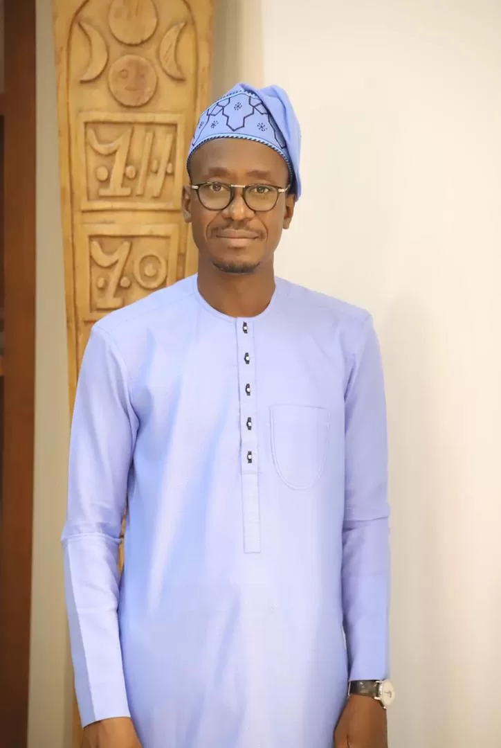 Homme souriant portant une tenue traditionnelle bleue et un bonnet assorti devant un mur clair et un panneau sculpté en bois