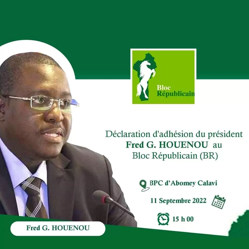 Homme en costume sombre et lunettes parlant devant un micro avec un logo vert Bloc Républicain en arrière-plan