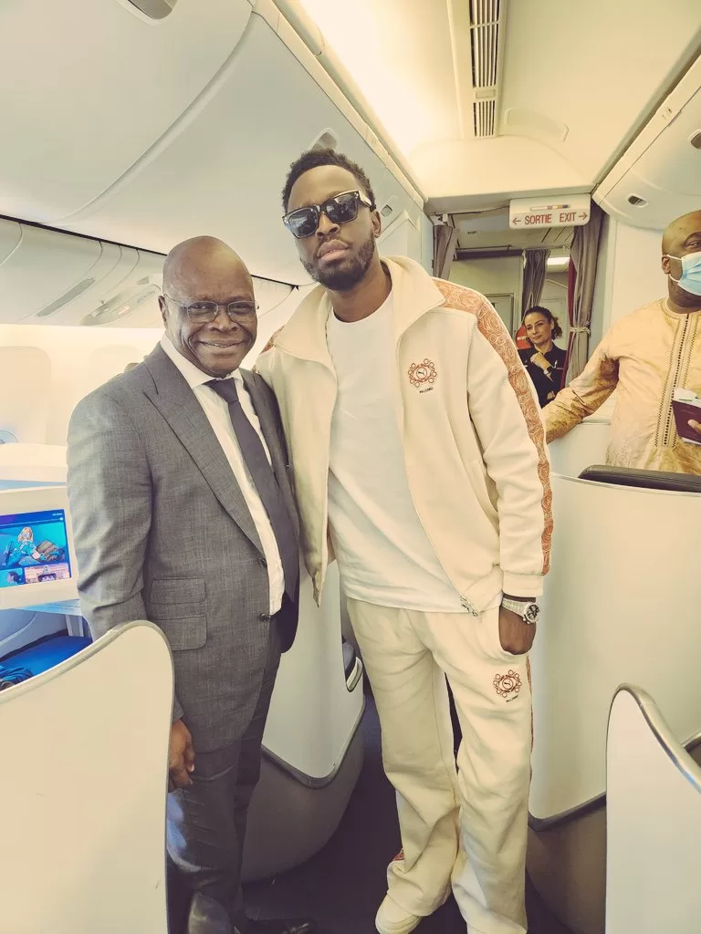 Deux hommes debout dans l'allée d'un avion, l'un en costume, l'autre en tenue décontractée avec lunettes de soleil, souriant