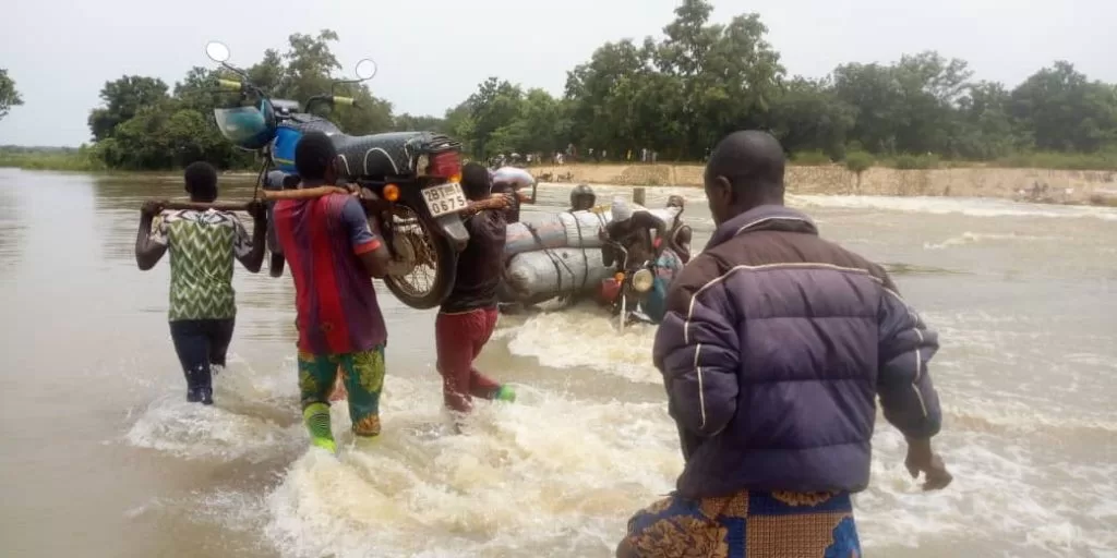 Quatre personnes portant des sacs et une moto traversent un cours d’eau en crue entouré d’arbres et de végétation