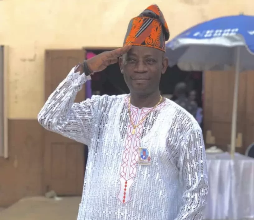 Un homme souriant en tenue traditionnelle blanche et chapeau orange fait un salut militaire devant un bâtiment beige.