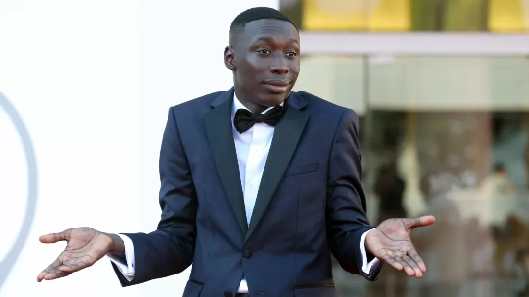 Un homme en costume noir avec un nœud papillon fait un geste d'incertitude avec les bras écartés et sourit légèrement