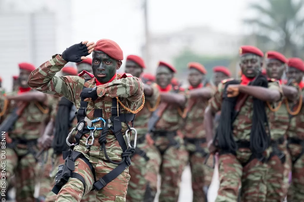 Soldats en uniforme camouflage et bérets rouges avec visage peint effectuant un salut militaire en formation.
