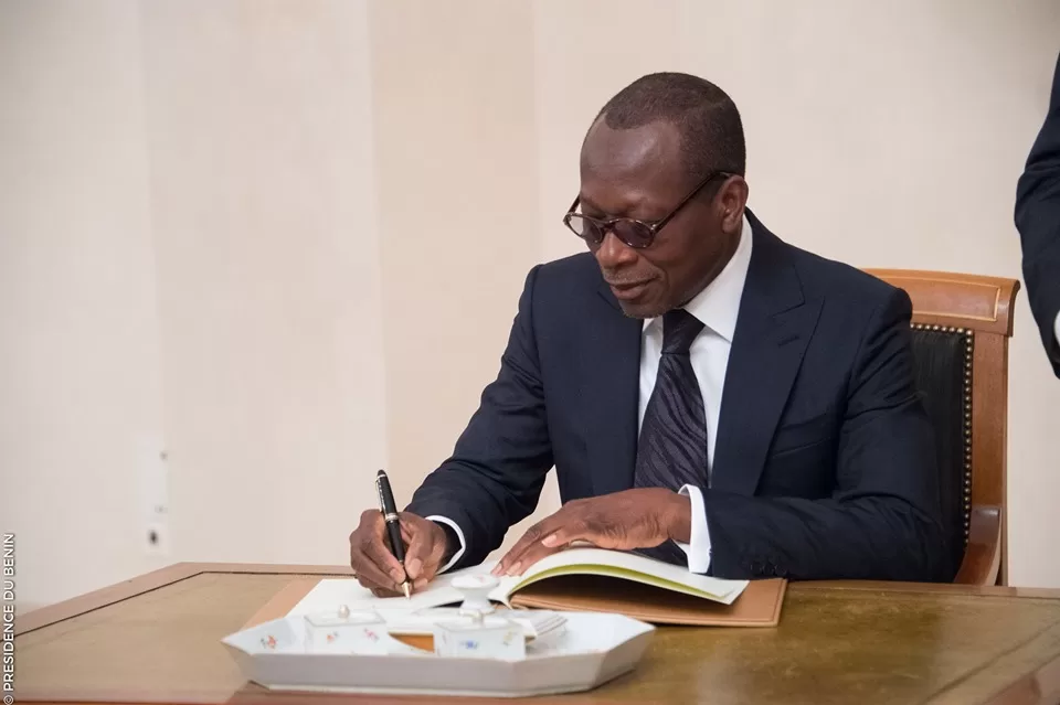 Un homme en costume sombre et lunettes signe un document posé sur une table en bois clair devant un mur beige