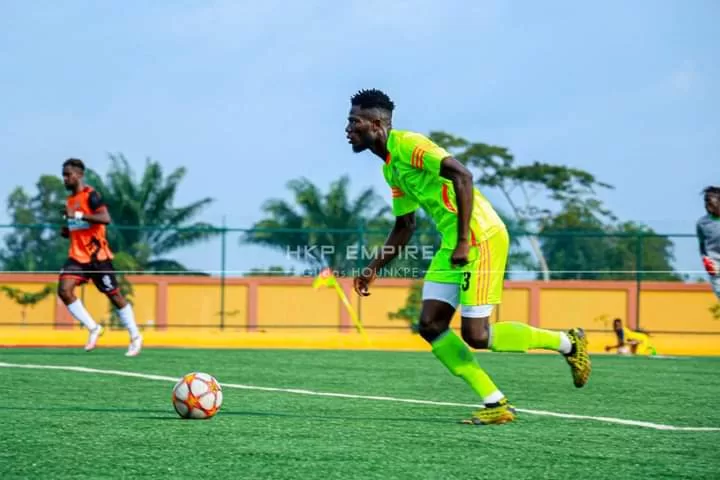 Joueur de football en tenue verte courant vers le ballon sur un terrain synthétique entouré d'autres joueurs en arrière-plan