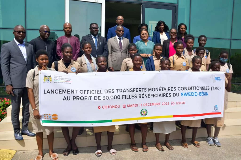Un groupe d’adultes et d’adolescentes posent devant un bâtiment avec une grande bannière annonçant un lancement officiel de transferts monétaires conditionnels