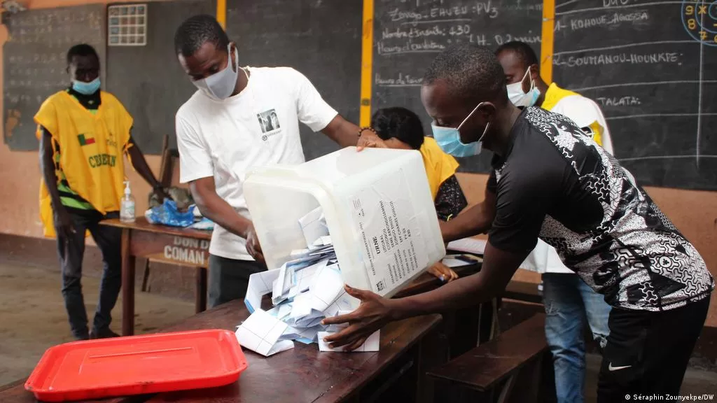 Des personnes en masque manipulent des bulletins de vote sortant d’une urne dans une salle de classe avec tableau noir en arrière-plan