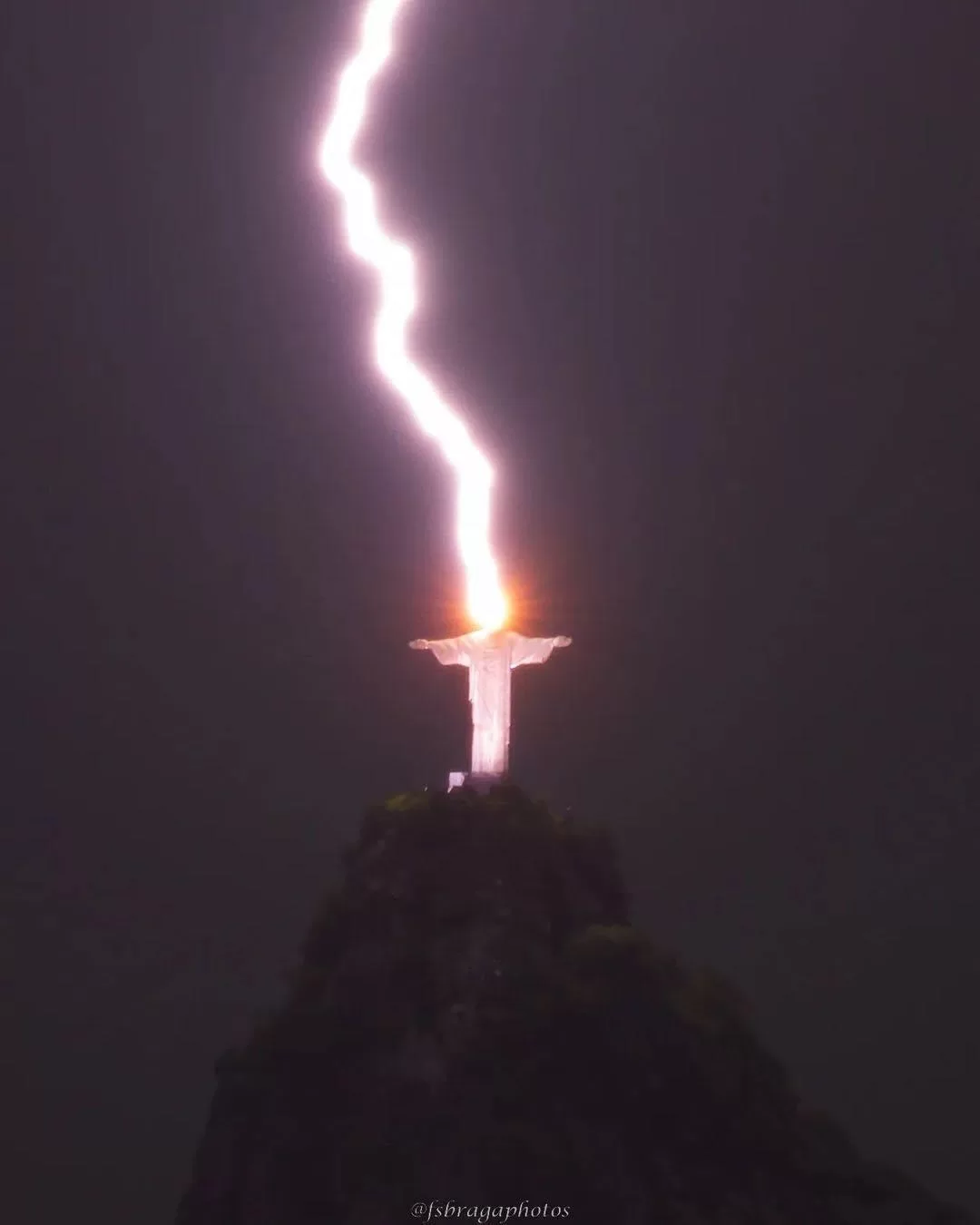 Éclair frappant simultanément une statue géante aux bras déployés sur un sommet rocheux dans la nuit