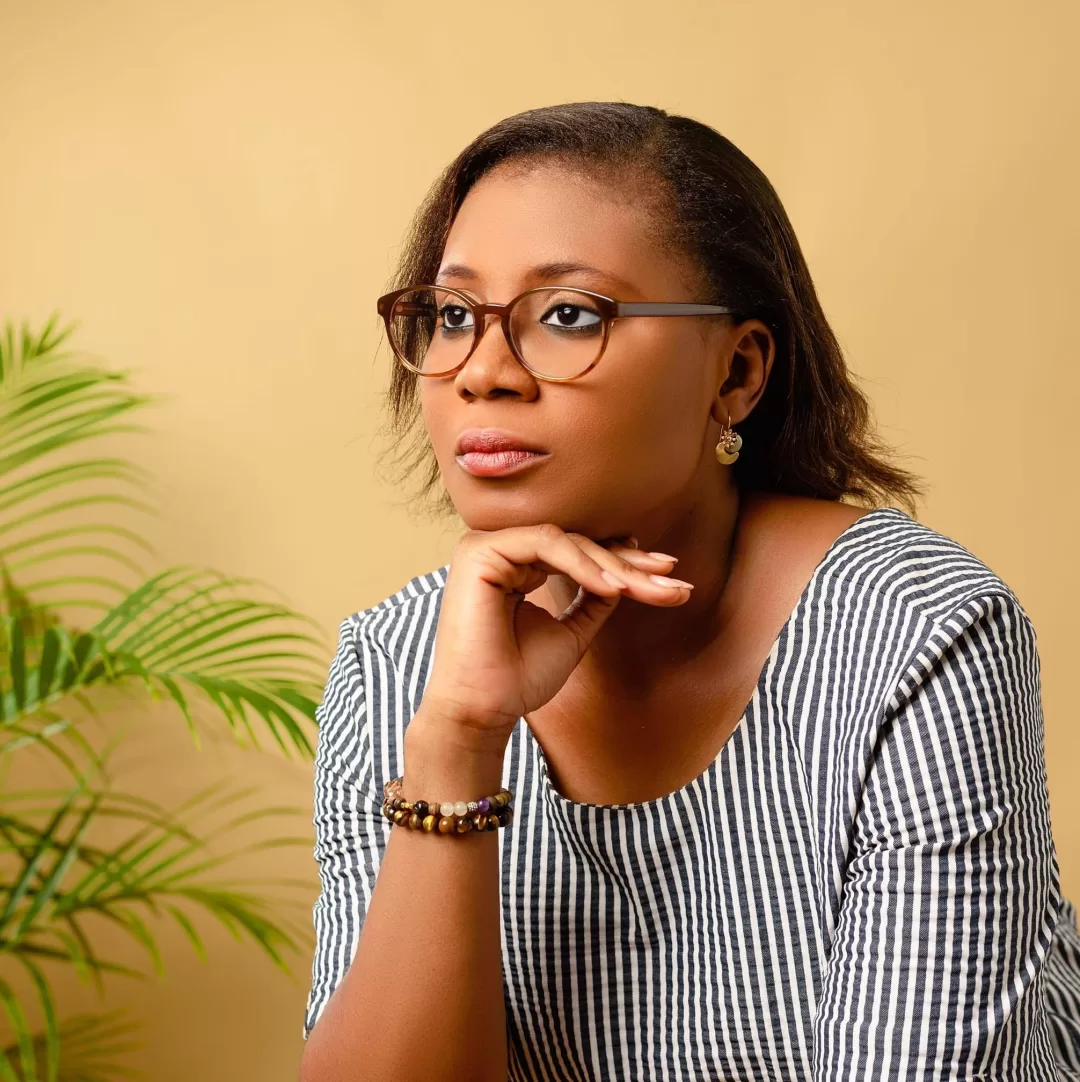 Femme portant des lunettes posant son menton sur la main avec un fond beige et une plante verte sur la gauche
